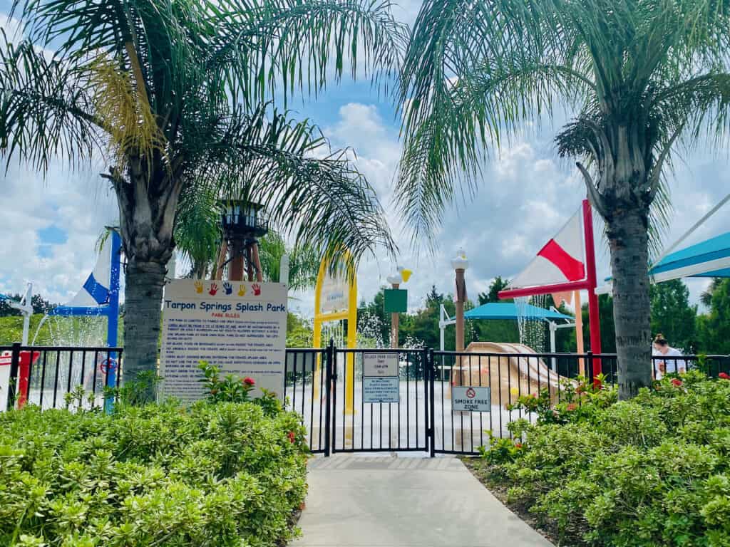Fenced gate area of the Tarpon Springs Splash Pad with water sprinklers in the distance.