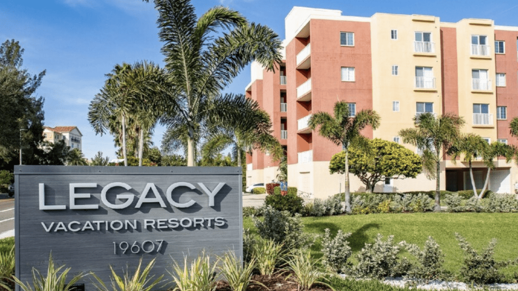 Legacy Vacation Resort Indian Shores exterior photo of the burnt orange and yellow exterior with palm trees near the water.